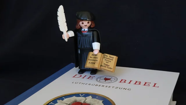 Die Darstellung zeigt eine Playmobil-Figur eines Reformators mit einer Feder und einer geöffneten Bibel auf einem Buch mit dem Titel "Die Bibel - Lutherübersetzung". Im Hintergrund ist ein symbolisches Wappen zu sehen, das eine rosa Blume mit einem Kreuz zeigt.