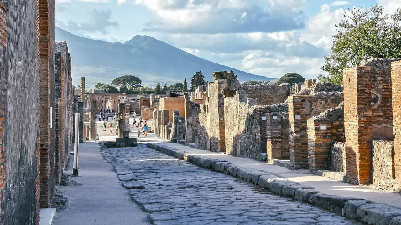 Blick auf eine antike Straße in Pompeji, umgeben von Ruinen. Im Hintergrund erhebt sich ein Berg, wahrscheinlich der Vesuv, unter einem blauen Himmel mit weißen Wolken.