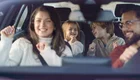 Eine Familie sitzt im Auto und hat Spaß. Die Mutter und die Kinder tanzen und lächeln, während der Vater am Steuer sitzt. Die Stimmung ist fröhlich und entspannt, was eine gemeinsame gute Zeit im Auto zeigt.