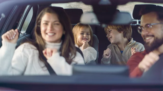 Eine Familie sitzt im Auto und hat Spaß. Die Mutter und die Kinder tanzen und lächeln, während der Vater am Steuer sitzt. Die Stimmung ist fröhlich und entspannt, was eine gemeinsame gute Zeit im Auto zeigt.