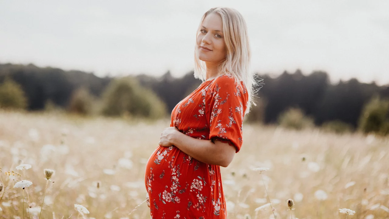 Eine schwangere Frau steht in einem blühenden Feld. Sie trägt ein rotes Kleid mit Blumenmuster und lächelt sanft. Ihre Hände legen sich auf ihren runden Bauch, während sie in die Kamera blickt. Der Hintergrund zeigt eine sanfte, grüne Landschaft.