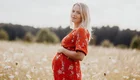 Eine schwangere Frau steht in einem blühenden Feld. Sie trägt ein rotes Kleid mit Blumenmuster und lächelt sanft. Ihre Hände legen sich auf ihren runden Bauch, während sie in die Kamera blickt. Der Hintergrund zeigt eine sanfte, grüne Landschaft.