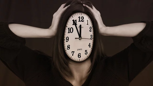 Eine Person mit einer Uhr als Gesicht hält sich die Hände am Kopf. Die Uhr zeigt die Zeit an, was ein Gefühl von Stress oder Zeitdruck vermittelt. Der Hintergrund ist in gedeckten Farben gehalten, was die emotionale Wirkung verstärkt.