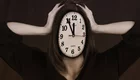 Eine Person mit einer Uhr als Gesicht hält sich die Hände am Kopf. Die Uhr zeigt die Zeit an, was ein Gefühl von Stress oder Zeitdruck vermittelt. Der Hintergrund ist in gedeckten Farben gehalten, was die emotionale Wirkung verstärkt.