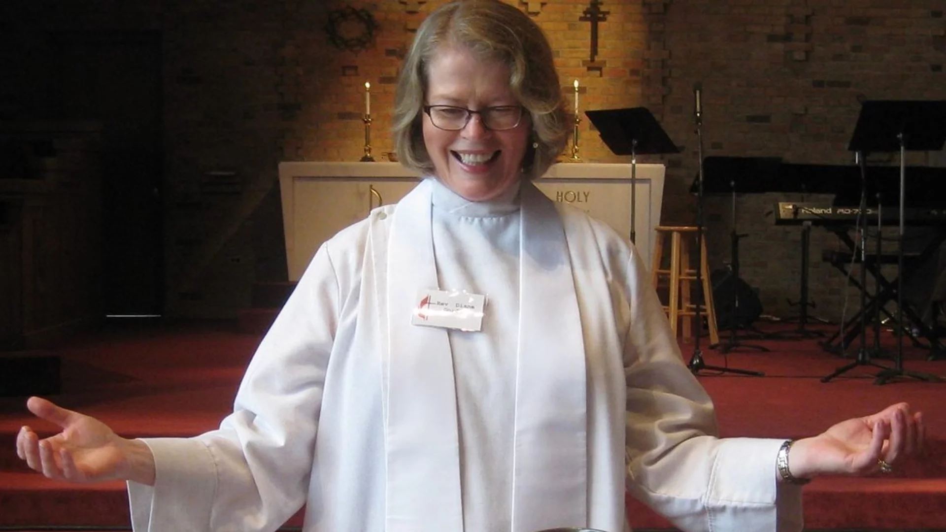 Eine lächelnde Frau in einem weißen Gewand steht mit ausgebreiteten Armen vor einem Altar in einer Kirche. Sie strahlt Freude und Offenheit aus, während sie eine geistliche Botschaft übermittelt. Der Hintergrund zeigt eine entspannte, kirchliche Umgebung mit Holz- und Lichtelementen.