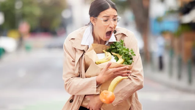 Eine Frau in einem beigen Mantel trägt eine Einkaufstasche mit Gemüse und Obst. Sie schaut überrascht oder erschrocken, während sie mit beiden Händen die Lebensmittel festhält. Die Szene wirkt lebhaft und vermittelt ein Gefühl von Unerwartetem.