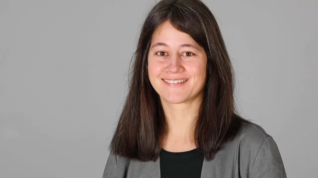 Eine Frau mit langen, dunklen Haaren lächelt in die Kamera. Sie trägt ein schwarzes T-Shirt und eine graue Jacke. Der Hintergrund ist neutral und hellgrau. Die Aufnahme wirkt freundlich und professionell.