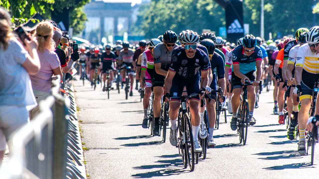 Eine große Gruppe von Radfahrern fährt mit hoher Geschwindigkeit unter einem Bogen mit der Aufschrift "START". Die Szene spielt sich auf einer breiten Straße ab, umgeben von Zuschauern und grünen Bäumen im Hintergrund. Es ist ein sonniger Tag.