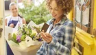 Eine Frau mit lockigem Haar steht an einer gelben Haustür und hält einen Blumenstrauß in der Hand. Sie lächelt und schaut auf eine Karte, die sie dabei hat. Im Hintergrund steht eine zweite Frau, die freundlich wirkt.