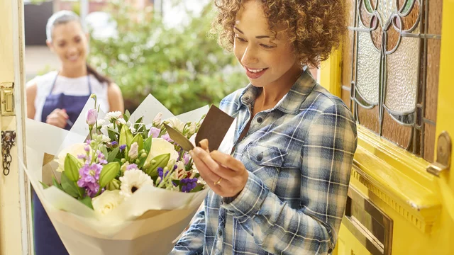 Eine Frau mit lockigem Haar steht an einer gelben Haustür und hält einen Blumenstrauß in der Hand. Sie lächelt und schaut auf eine Karte, die sie dabei hat. Im Hintergrund steht eine zweite Frau, die freundlich wirkt.