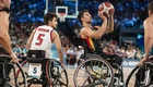 Ein Basketballspiel im Rollstuhl, bei dem ein Spieler in gelber Uniform den Ball wirft. Ein anderer Spieler in roter Uniform beobachtet ihn aufmerksam. Im Hintergrund sind Zuschauer und die Spielfeldmarkierungen sichtbar.