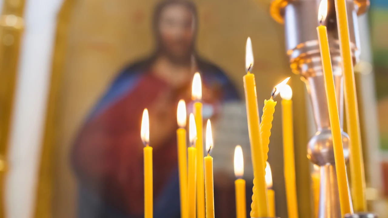 Einige brennende Kerzen stehen auf einem Tisch. Im Hintergrund ist eine unscharfe religiöse Ikone sichtbar, die eine Figur darstellt. Die Kerzen sind in unterschiedlichen Höhen und leuchten warm in einer orthodoxen Kirche. 