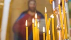 Einige brennende Kerzen stehen auf einem Tisch. Im Hintergrund ist eine unscharfe religiöse Ikone sichtbar, die eine Figur darstellt. Die Kerzen sind in unterschiedlichen Höhen und leuchten warm in einer orthodoxen Kirche. 