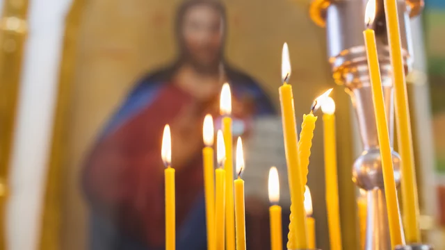 Einige brennende Kerzen stehen auf einem Tisch. Im Hintergrund ist eine unscharfe religiöse Ikone sichtbar, die eine Figur darstellt. Die Kerzen sind in unterschiedlichen Höhen und leuchten warm in einer orthodoxen Kirche. 