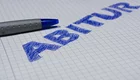 Auf einem karierten Blatt Papier steht in blauer Schrift das Wort "ABITUR". Neben dem Wort liegt ein grauer Kugelschreiber.