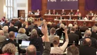 Eine Versammlung mit vielen Teilnehmern ist zu sehen, die an Tischen sitzen und auf einen Redner blickten. Eine Person in der Mitte hat die Hand erhoben, um eine Frage zu stellen. Im Hintergrund sitzen weitere Redner. Ein Banner kündigt die 8. Tagung an.