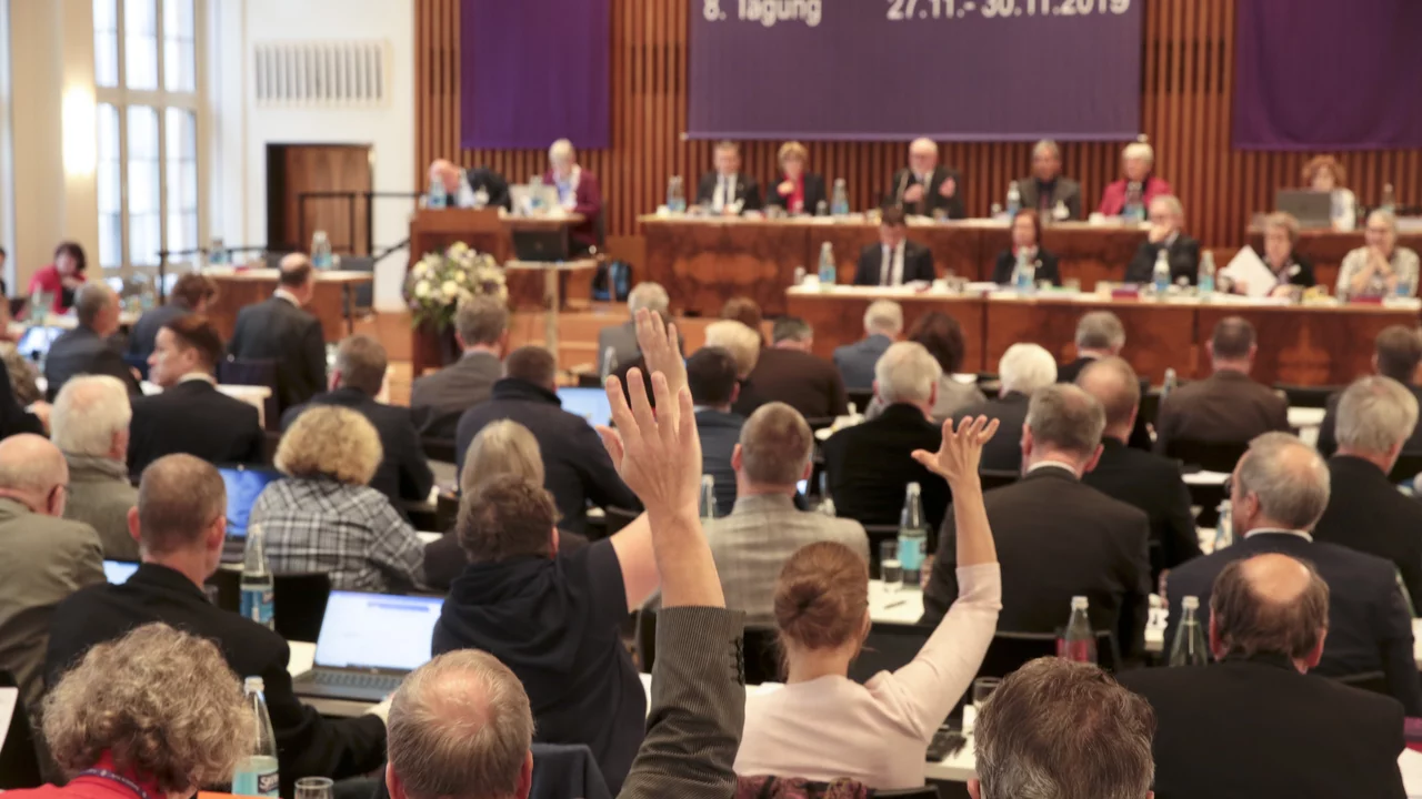 Eine Versammlung mit vielen Teilnehmern ist zu sehen, die an Tischen sitzen und auf einen Redner blickten. Eine Person in der Mitte hat die Hand erhoben, um eine Frage zu stellen. Im Hintergrund sitzen weitere Redner. Ein Banner kündigt die 8. Tagung an.