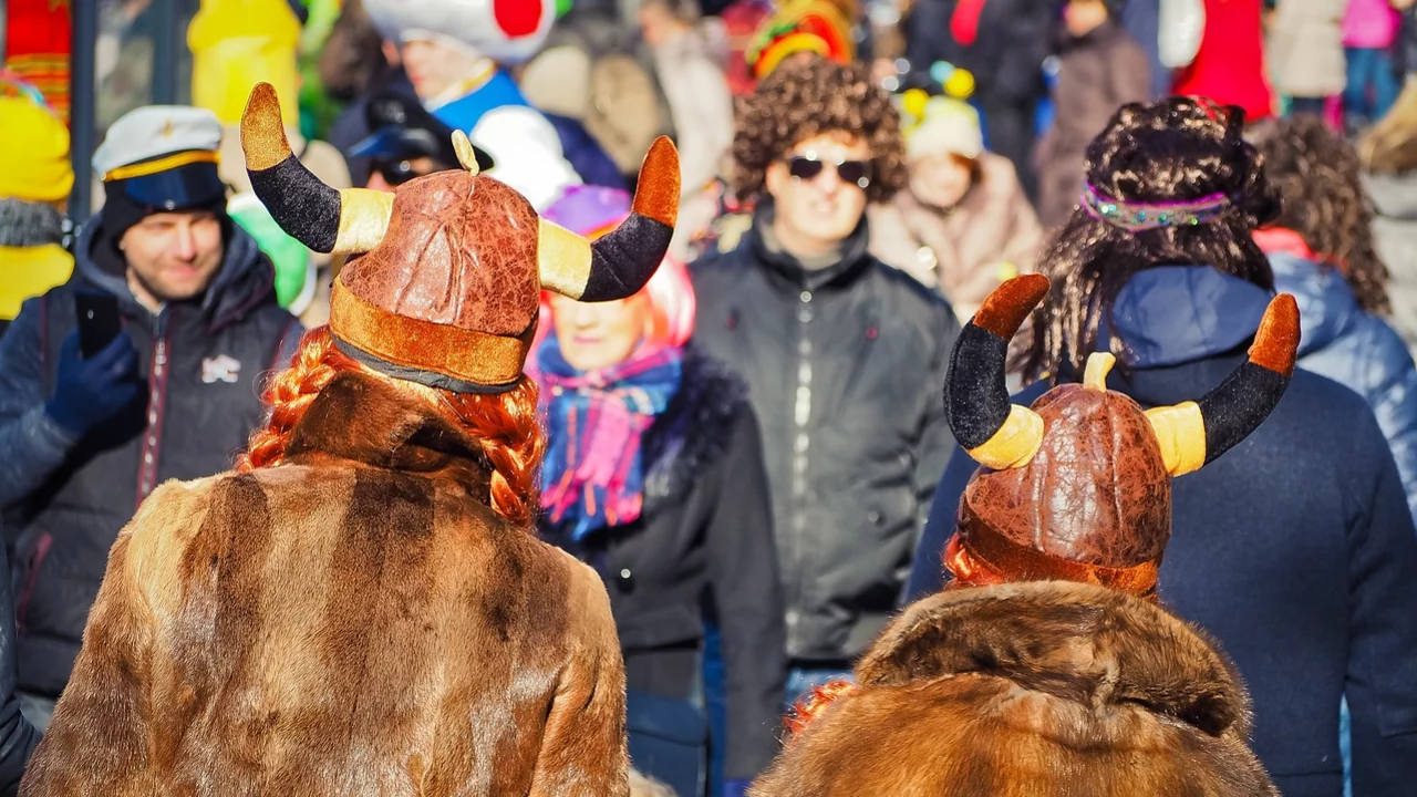 Zwei Personen mit gehörnten Vikingermützen und Fellen stehen Rücken an Rücken in einer belebten Menschenmenge. Im Hintergrund sind viele bunte Kostüme und Faschingsfeiern zu erkennen, was auf eine festliche Atmosphäre hinweist.