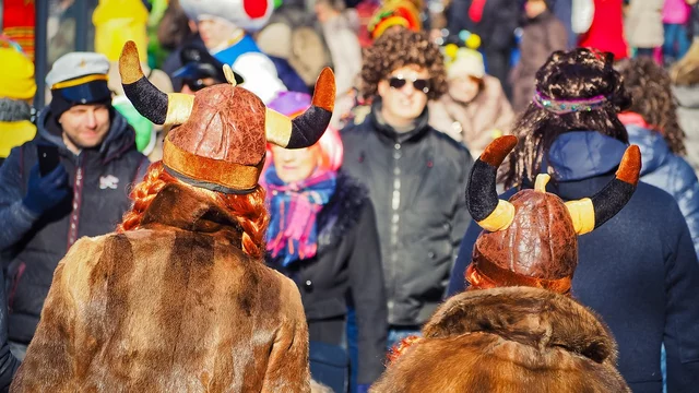 Zwei Personen mit gehörnten Vikingermützen und Fellen stehen Rücken an Rücken in einer belebten Menschenmenge. Im Hintergrund sind viele bunte Kostüme und Faschingsfeiern zu erkennen, was auf eine festliche Atmosphäre hinweist.