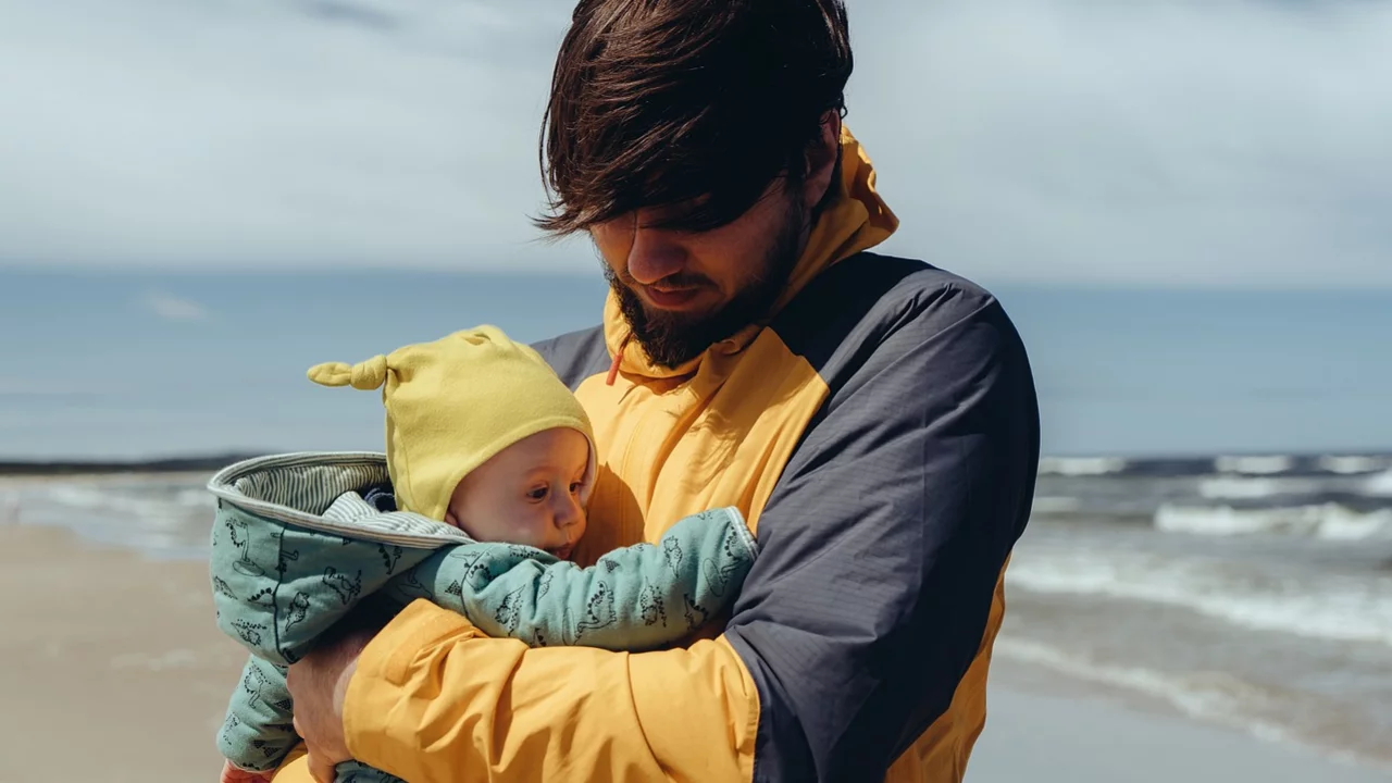 Ein Mann mit kurzem Haar hält ein Baby in einem grünen Mantel auf dem Arm. Beide schauen miteinander, während sie am Strand stehen. Im Hintergrund sind das Meer und der Himmel sichtbar. Der Mann trägt eine gelbe und graue Jacke.