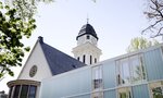 Das Bild zeigt eine hybride Architektur mit einem historischen Gebäude auf der linken Seite, das eine gichtige Fassade und einen Turm mit Ziffernblatt hat, und einem modernen, gläsernen Anbau auf der rechten Seite, umgeben von Bäumen und einem blauen Himmel.