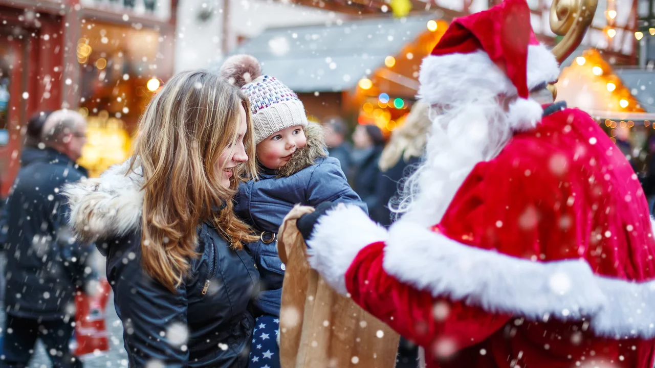 Eine Frau hält ein Kind in den Armen, während sie mit dem Weihnachtsmann spricht. Das Kind trägt eine Mütze und schaut neugierig. Es fällt Schnee, und die Umgebung wirkt festlich mit beleuchteten Ständen.