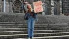 Eine Person steht auf einer Treppe vor einer beeindruckenden gotischen Architektur. Sie hält ein Schild mit der Aufschrift „TELL ME YOUR GOOD NEWS“. Die Person trägt eine Jeansjacke und blickt lächelnd in die Kamera.