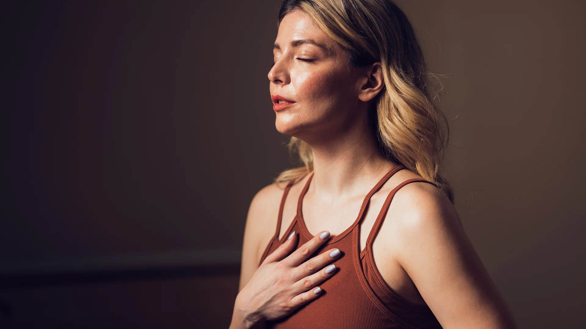 Eine Frau mit langen, lockigen Haaren trägt ein braunes Oberteil. Sie steht mit geschlossenen Augen und hand auf ihrer Brust, während sie entspannt wirkt. Der Hintergrund ist einfach und dunkel, was die ruhige Atmosphäre verstärkt.