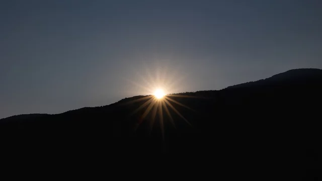 Eine aufgehende Sonne strahlt hinter einem Hügel hervor, umgeben von einem dunklen Himmel. Die Sonnenstrahlen erzeugen ein schönes Lichtspiel, das die Silhouette der Berge umreißt.