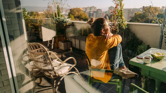 Eine Frau sitzt mit angezogenen Beinen auf einem Stuhl auf dem Balkon, der mit Pflanzen und Balkonmöbeln zugestellt ist. Sie hat die Arme um die Knie gelegt und schaut in die Sonne. Das Bild strahlt eine gemütliche Atmosphäre aus.