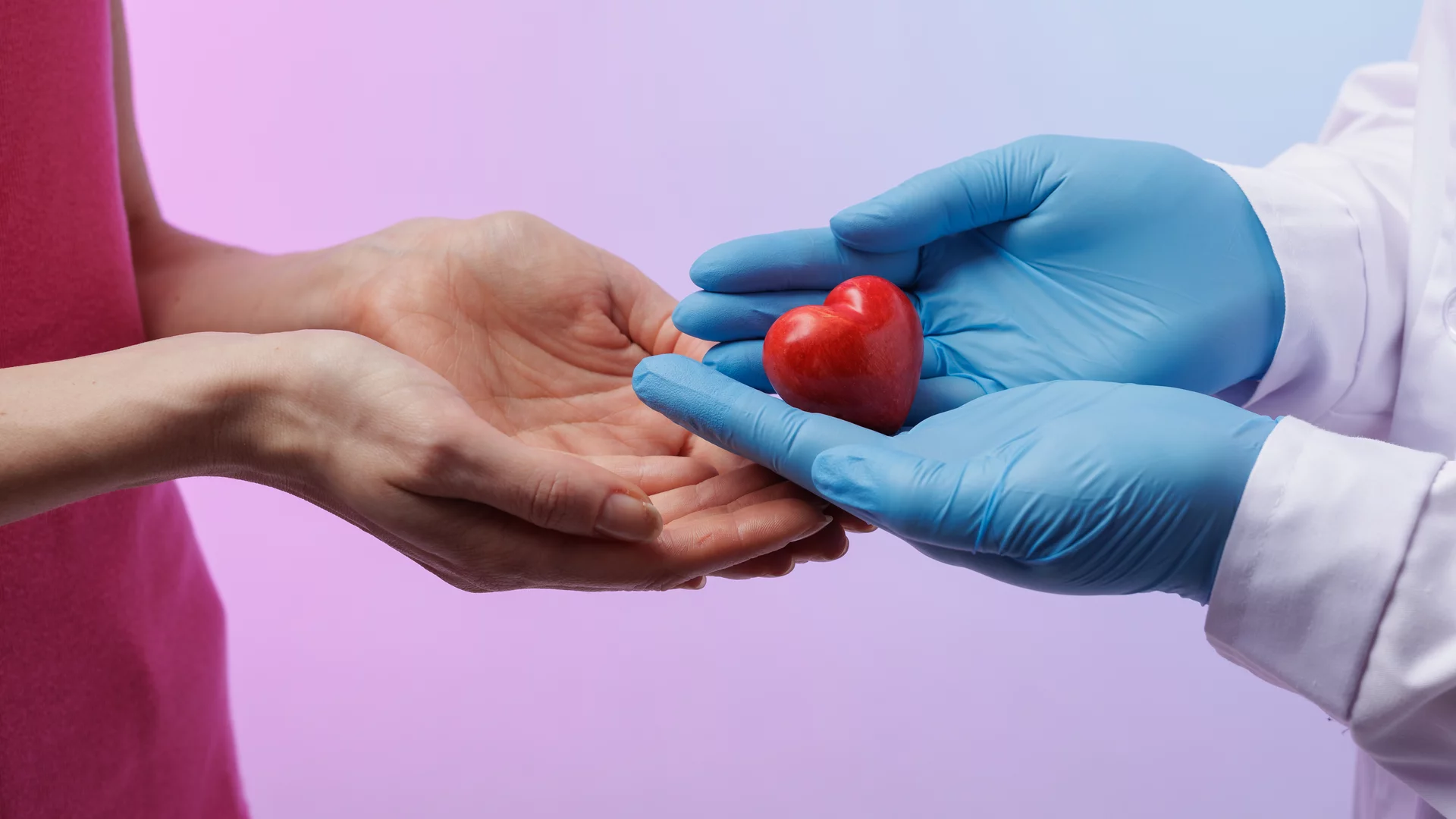 Eine Hand in blauen Handschuhen hält ein rotes Herz, während eine zweite Hand bereit ist, es zu empfangen. Der Hintergrund ist in sanften Lila- und Rosa-Tönen gehalten. Das Bild symbolisiert Fürsorge und den Austausch von Liebe oder Unterstützung.