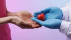 Eine Hand in blauen Handschuhen hält ein rotes Herz, während eine zweite Hand bereit ist, es zu empfangen. Der Hintergrund ist in sanften Lila- und Rosa-Tönen gehalten. Das Bild symbolisiert Fürsorge und den Austausch von Liebe oder Unterstützung.