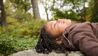Ein Mädchen mit lockigem Haar liegt entspannt auf einem Stein und blickt zur Decke. Um sie herum sind grüne Pflanzen und Bäume, die eine ruhige, natürliche Umgebung schaffen. Die Szene vermittelt ein Gefühl von Frieden und Entspannung in der Natur.