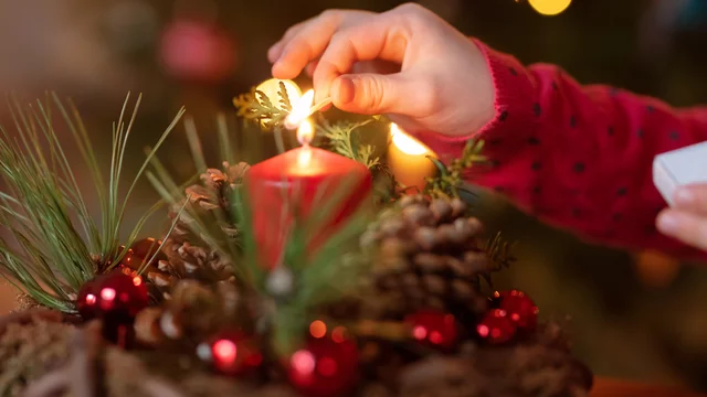 Eine Hand zündet mit einem Streichholz eine rote Kerze in einem Adventsgesteck an, das aus Kiefernzweigen, roten Kugeln und  Kiefernzweigen besteht.