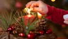 Eine Hand zündet mit einem Streichholz eine rote Kerze in einem Adventsgesteck an, das aus Kiefernzweigen, roten Kugeln und  Kiefernzweigen besteht.