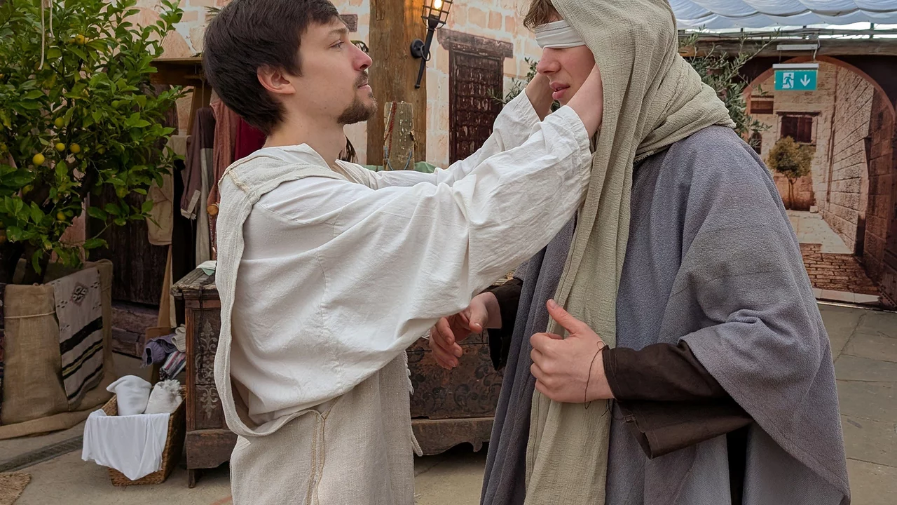 Ein Mann in einem hellen Gewand nimmt einem anderen Mann, der ein graues Gewand trägt, die Augenbinde ab. Die Szene spielt in einer historischen Kulisse mit verschiedenen Requisiten und Pflanzen im Hintergrund.