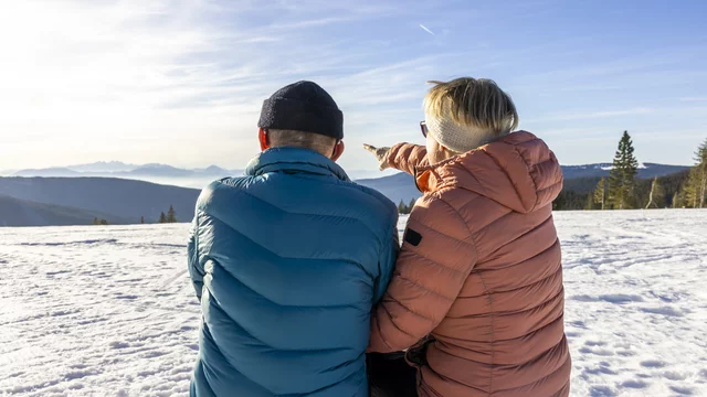 Zwei Personen sitzen auf einer Holzbank im Schnee und schauen auf eine hügelige Landschaft. Eine Person weist mit dem Finger in die Ferne, während die Sonne scheint und die Berge im Hintergrund erkennbar sind. Es vermittelt eine friedliche Winteratmosphäre und Mut zu Neuem. 