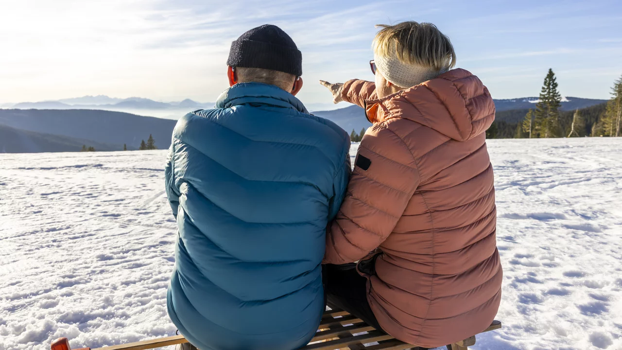 Zwei Personen sitzen auf einer Holzbank im Schnee und schauen auf eine hügelige Landschaft. Eine Person weist mit dem Finger in die Ferne, während die Sonne scheint und die Berge im Hintergrund erkennbar sind. Es vermittelt eine friedliche Winteratmosphäre und Mut zu Neuem. 