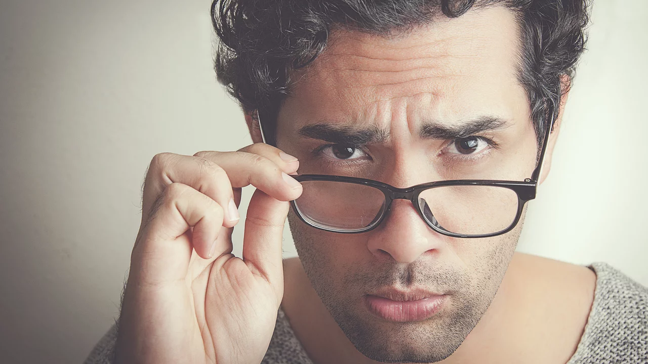 Ein junger Mann mit lockigem Haar schaut direkt in die Kamera, während er seine Brille anhebt. Sein Gesichtsausdruck ist ernst und nachdenklich, was ihm einen aufmerksamen und eindringlichen Blick verleiht.