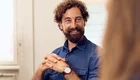 Ein lächelnder Mann mit lockigem Haar und Bart sitzt in einem modernen Raum. Er trägt ein blaues Hemd und hat die Hände entspannt vor sich gefaltet. Im Hintergrund sind verschwommene Farben zu sehen, die auf eine angenehme Atmosphäre hinweisen.