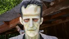 Eine lebensgroße Statue eines Frankenstein-ähnlichen Monsters mit blassem Gesicht, sichtbaren Narben und einem ernsthaften Ausdruck. Es trägt einen dunklen Anzug und hat eine Steckdose an der Halsregion. Die Statue steht vor einer Holzstruktur und ist von grünem Umland umgeben.
