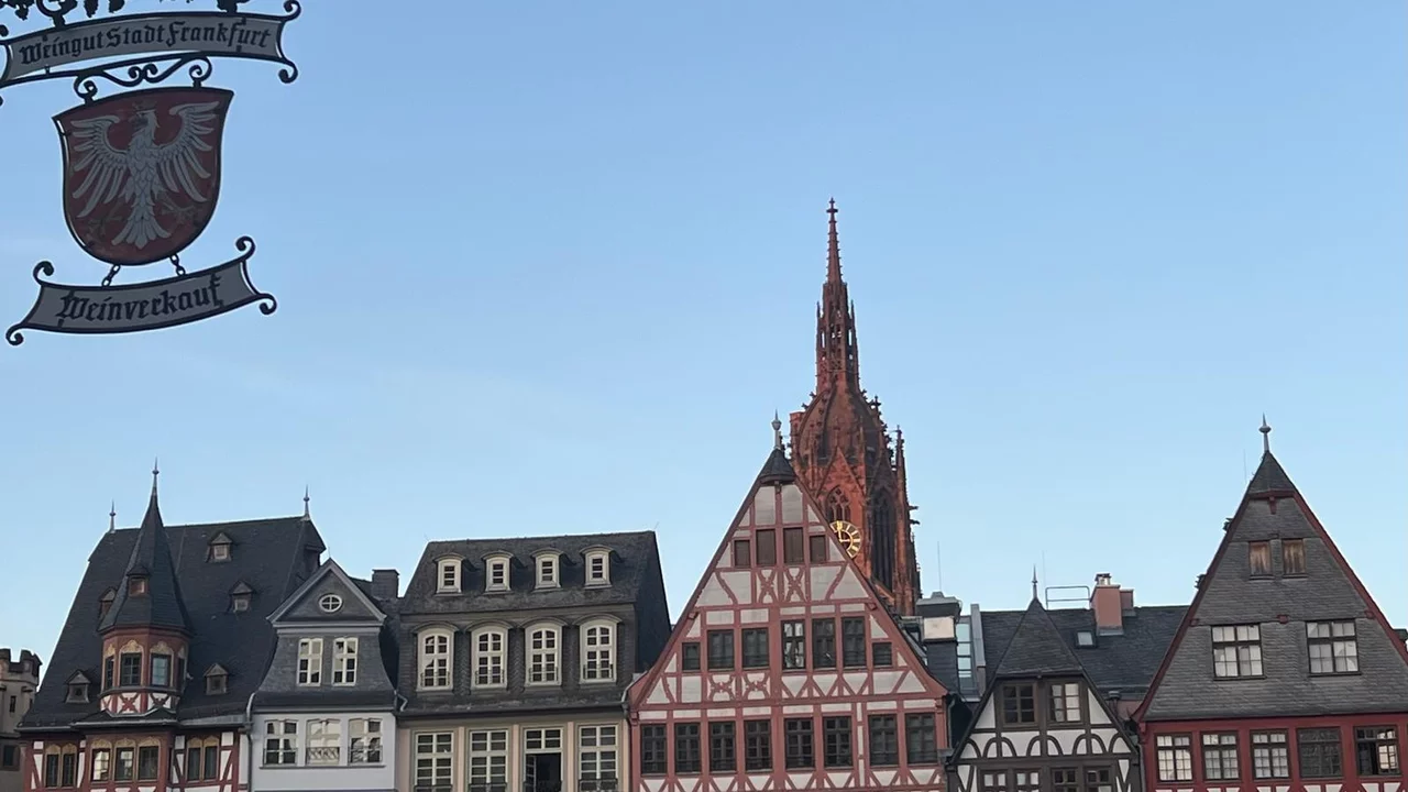 Ein historisches Stadtbild mit bunten Fachwerkhäusern in Frankfurt. Im Hintergrund ragt der Dom mit seinem markanten Turm in den Himmel. Der Himmel ist klar und blau, was die Architektur der Gebäude betont.