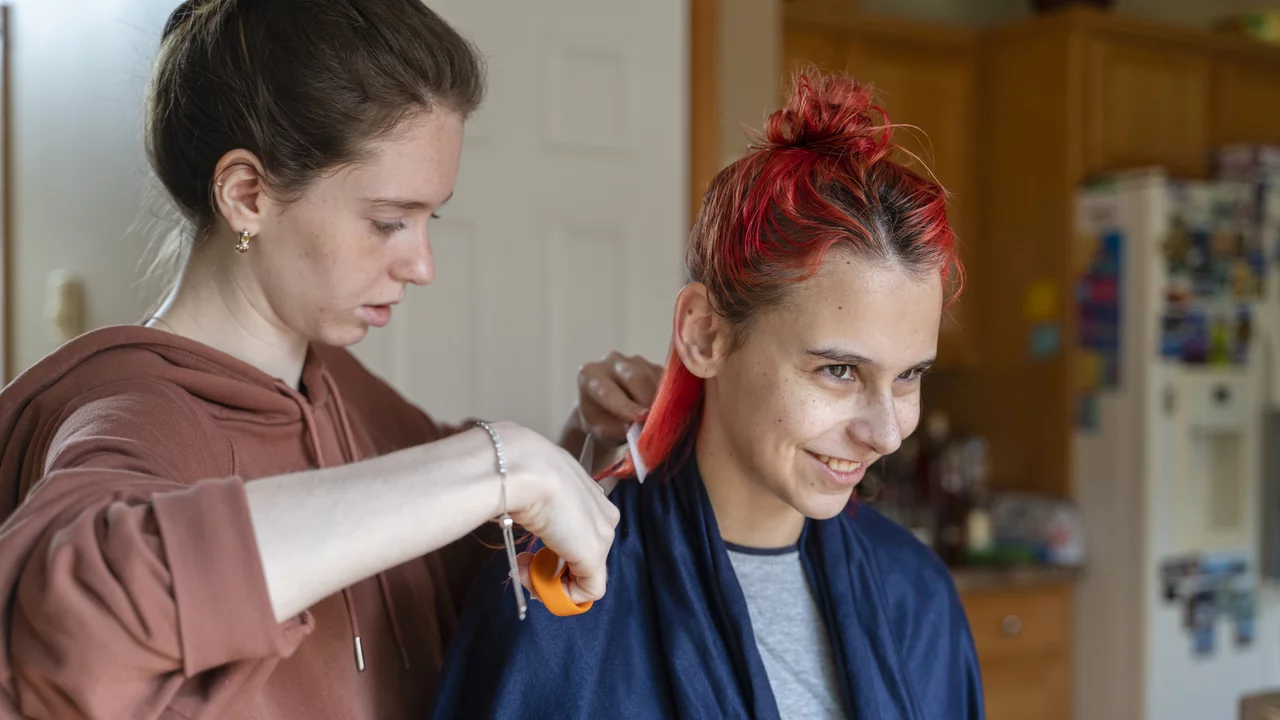 Eine Frau schneidet mit einer Schere das Haar einer anderen Frau, die ein zufriedenes Lächeln zeigt. Die Frisur wird gerade bearbeitet, während die Haarenden sichtbar rot gefärbt sind. Beide Frauen tragen bequeme Kleidung und stehen in einer häuslichen Umgebung.