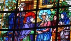 Das Bild zeigt ein buntes Kirchenfenster mit biblischen Figuren. Eine weibliche Gestalt mit einem blauen Gewand spricht mit einer anderen Frau. Im Hintergrund stehen mehrere Personen und eine Esel. Der Szene sind Bäume und ein Gebäude hinzugefügt, die eine religiöse Atmosphäre schaffen.
