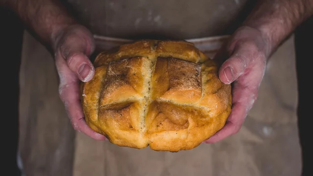 Eine Person hält ein frisch gebackenes Brot in den Händen. Das Brot hat eine goldene Kruste mit einer kreuzförmigen Einschnittmarkierung. Die Hände der Person sind mit Mehl bestaubt, was auf das Bäckerhandwerk hindeutet.