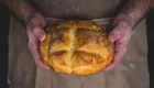 Eine Person hält ein frisch gebackenes Brot in den Händen. Das Brot hat eine goldene Kruste mit einer kreuzförmigen Einschnittmarkierung. Die Hände der Person sind mit Mehl bestaubt, was auf das Bäckerhandwerk hindeutet.