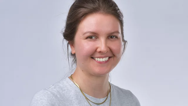 Eine junge Frau lächelt freundlich und steht vor einem neutralen Hintergrund. Sie trägt ein graues T-Shirt und eine goldene Kette. Ihr Haar ist locker zurückgebunden. Die Bildkomposition hebt ihren freundlichen Gesichtsausdruck hervor.