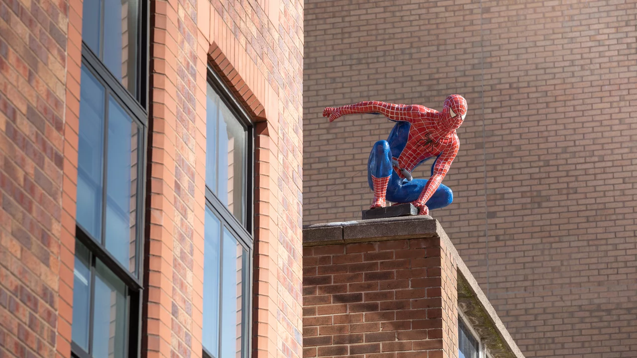 Eine Spider-Man-Figur sitzt auf dem Dach eines Gebäudes, mit einem dynamischen Pose, als ob sie bereit ist, in die Stadt zu springen. Die Figur hat ein rot-blaues Kostüm mit dem typischen Spinnennetz-Muster. Im Hintergrund sind mehrere Fenster des Gebäudes sichtbar.