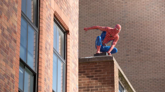 Eine Spider-Man-Figur sitzt auf dem Dach eines Gebäudes, mit einem dynamischen Pose, als ob sie bereit ist, in die Stadt zu springen. Die Figur hat ein rot-blaues Kostüm mit dem typischen Spinnennetz-Muster. Im Hintergrund sind mehrere Fenster des Gebäudes sichtbar.