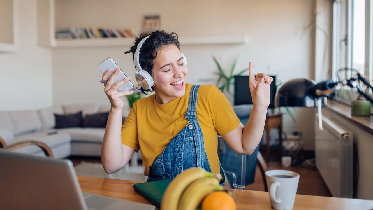 Eine junge Frau mit Kopfhörern sitzt an einem Tisch und hört Musik. Sie hält ihr Smartphone in der Hand und lächelt, während sie zur Musik tanzt. Auf dem Tisch liegen eine Tasse Kaffee und einige Früchte. Der Hintergrund zeigt einen hellen Raum mit Pflanzen und einem gemütlichen Sofa.